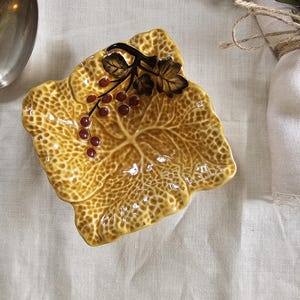 Puede incluir: Un plato decorativo de cerámica con forma de hoja, con una superficie texturizada de color amarillo-marrón. El plato está adornado con una rama marrón oscuro con hojas y racimos de bayas rojas. Una cuchara plateada y una ramita de romero están en el fondo.