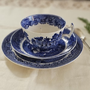 May include: A blue and white Willow pattern teacup and saucer set. The teacup sits on a matching saucer and a larger plate. The set features a detailed landscape design in a classic blue and white colour scheme, ideal for afternoon tea.