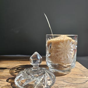 Op de afbeelding: Een heldere, geslepen glazen suikerpot met een deksel en een bijpassend glas gevuld met bruine suiker. Een veer is in de suiker geplaatst. De kom en het glas staan op een houten oppervlak.