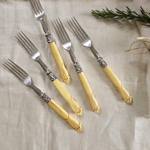May include: A set of antique forks with silver tines and cream-coloured handles. The handles have a decorative design. The forks are arranged on a neutral linen surface, suggesting a dining or table setting. The forks are likely made of silver.