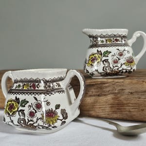 Könnte beinhalten: Zwei antike Sahnekännchen mit floralen Mustern. Die Kännchen sind weiß mit braunen, gelben und roten floralen Designs. Eines hat einen Ausguss und einen Griff, das andere hat zwei Griffe. Ein silberner Löffel liegt auf einer weißen Oberfläche.