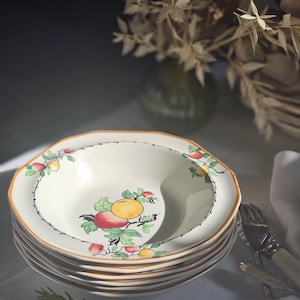 May include: A stack of cream-coloured ceramic bowls with a scalloped edge and a fruit design. The bowls have a gold rim and a colourful illustration of red and yellow fruit with green leaves. The bowls are stacked on a white tablecloth.
