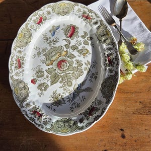 Może przedstawiać: Owalny półmisek ceramiczny z kremową podstawą i szczegółowym wzorem kwiatowym w odcieniach zieleni, czerwieni i żółci, podkreślony motywem ptaka i motyla. Srebrny widelec i łyżka spoczywają na białej serwetce obok półmiska.