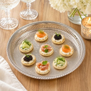 May include: A round silver serving tray with an ornate pierced rim and engraved centre, holding seven round crackers topped with various ingredients including smoked salmon, black caviar, sliced cucumber, and cream cheese, garnished with fresh herbs.