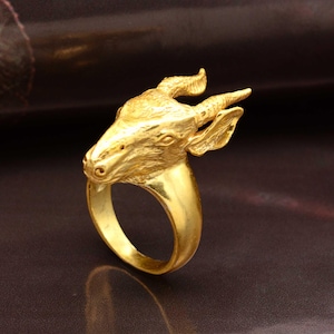 Może przedstawiać: Złoty pierścionek z szczegółową rzeźbą głowy gazeli. Pierścionek ma gładką, wypolerowaną obrączkę, a głowa gazeli jest misternie zaprojektowana z rogami i rysami twarzy. Pierścionek jest na ciemnym tle.
