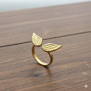 Puede incluir: Un anillo dorado con diseño de alas. El anillo tiene dos alas detalladas, una a cada lado, creando un diseño abierto. El anillo está sobre una superficie de madera.