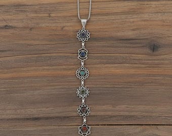 May include: A silver-toned necklace featuring seven chakra symbols, each with a different colored gemstone. The pendant hangs from a delicate chain against a wooden background. The gemstones include shades of purple, blue, green, yellow, red, and orange.