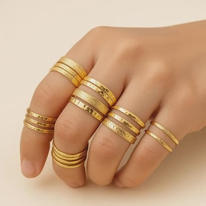 May include: A hand adorned with several gold rings. The rings feature diverse designs, such as plain bands, textured bands, and stacked bands. All the rings are gold in colour and seem to be made of metal.