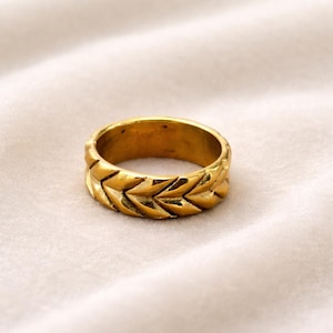 Puede incluir: Un anillo dorado con un patrón de chevrón repetido. El anillo es circular y parece ser de metal. El anillo está sobre un fondo blanco.