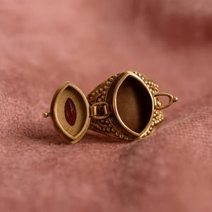 May include: A detailed, open locket ring in antique gold. The ring features an oval compartment with a small, red gemstone inside. The ring is set against a pink background, highlighting the intricate design and craftsmanship.