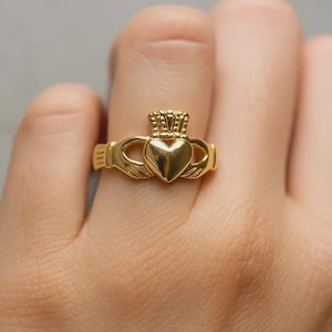 May include: A gold-colored Claddagh ring featuring a heart, crown, and hands design. The ring is a symbol of love, loyalty, and friendship. The heart is in the center, topped with a crown, and held by two hands.