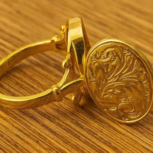 Op de afbeelding: Een gouden ring met een ovale, gegraveerde voorkant. De ring is gemaakt van goud en heeft een sierlijk ontwerp. De ring ligt op een houten oppervlak.