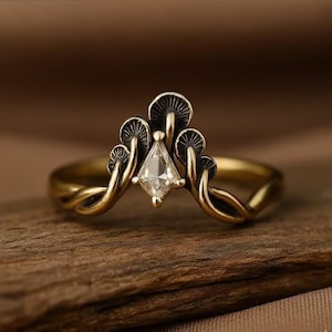 Può includere: Un anello dorato con una gemma trasparente a forma di diamante al centro. Il design dell'anello incorpora dettagli a forma di fungo, con una fascia che si curva attorno al dito. L'anello è appoggiato su una superficie di legno.