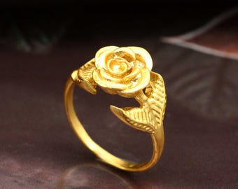 Può includere: Un anello color oro con un design a rosa dettagliato. La fascia dell'anello è liscia e la rosa è circondata da due foglie. L'anello è realizzato in un materiale metallico lucido.