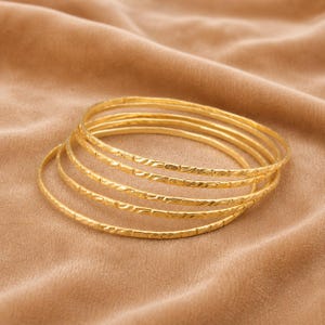 Può includere: Un set di cinque bracciali rigidi circolari e sottili color oro. Ogni bracciale presenta un motivo inciso sottile e ripetitivo lungo l'intera superficie esterna. I bracciali sono impilati in modo leggermente sfalsato.