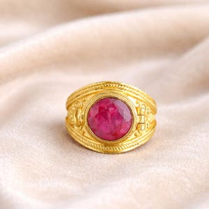 Op de afbeelding: Een gouden ring met een grote, ovale, gefacetteerde, robijnkleurige steen. De ring heeft ingewikkelde, in reliëf aangebrachte details rond de band en de zetting van de steen. De ring staat tegen een zachte, neutrale achtergrond.