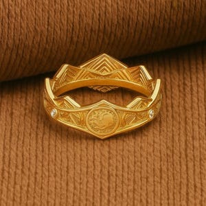 Könnte beinhalten: Ein goldfarbener Ring mit einem aufwendigen geometrischen Design. Der Ring hat ein zentrales kreisförmiges Emblem mit einer detaillierten Figur und zwei kleinen, klaren Edelsteinen. Der Ring ist vor einem strukturierten braunen Hintergrund platziert.