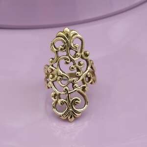 Könnte beinhalten: Ein goldfarbener Filigranring mit einem verzierten, durchbrochenen Design. Der Ring hat eine zentrale, längliche Form mit wirbelnden Mustern und floralen Details. Der Ring ist vor einem weichen, lila Hintergrund platziert.
