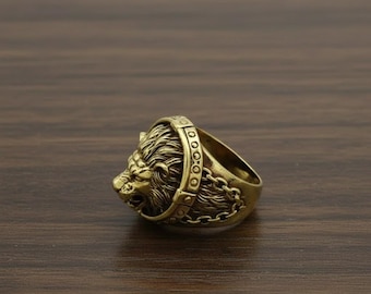 May include: A detailed, antique-style ring in a gold tone, featuring a lion's head with a textured mane and a chain detail around the ring's band. The ring is set against a dark wood grain surface.