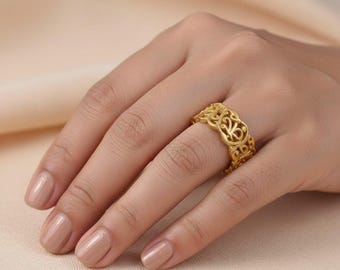 Puede incluir: Un anillo dorado con un intrincado diseño calado se lleva en un dedo. El patrón del anillo presenta formas arremolinadas y florales. El anillo se muestra en una mano con uñas pulidas, sobre un fondo neutro.
