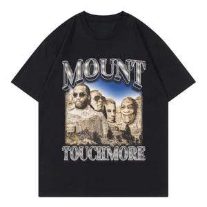 Könnte beinhalten: Ein schwarzes Kurzarm-T-Shirt mit einem Grafikdruck von vier in einen Berg gehauenen Gesichtern, die an den Mount Rushmore erinnern. Der Text MOUNT steht über der Grafik und TOUCHMORE darunter in einer metallisch silbernen Schrift.