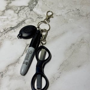 May include: A collection of everyday carry items on a key ring. Includes a black pen, small black scissors, a black LED light, and a metal key ring with two silver-colored clasps.