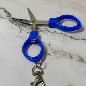 May include: A pair of small, open scissors with blue handles and a silver-colored blade. The scissors are attached to a silver-colored keychain clip. The scissors are on a white and gray marble surface.