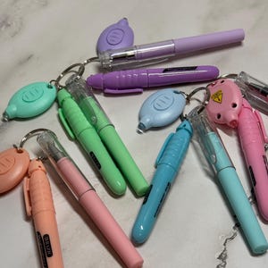 Puede incluir: Una colección de bolígrafos y llaveros coloridos en varios tonos pastel. Los bolígrafos son en tonos rosa, verde, azul y morado, con tapas transparentes. Los llaveros son de colores a juego y tienen forma ovalada.