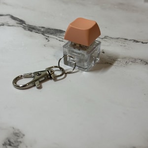 Puede incluir: Un llavero con una tecla de teclado. La tecla es de color melocotón y se asienta sobre una carcasa cuadrada transparente. Un cierre de color plateado está unido a la carcasa de la tecla con un pequeño anillo de metal. El fondo es una superficie con vetas de mármol.