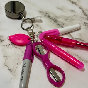 Può includere: Una collezione di forniture per ufficio rosa e argento su una superficie di marmo. Include un portabadge retrattile, una luce LED rosa, un pennarello Sharpie Mini viola, un paio di forbici viola e due penne rosa.