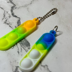 Puede incluir: Dos coloridos juguetes fidget con forma de vaina de guisantes. Uno es amarillo, verde y naranja, el otro es azul, verde y blanco. Cada uno tiene tres secciones con forma de burbuja. Ambos tienen una cadena o anillo de metal para sujetar.