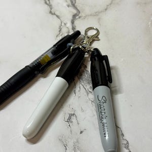 May include: A keychain with three writing instruments: a black Pilot pen, and two Sharpie Mini markers. One Sharpie is black and white, the other is gray with the brand name printed on the side. The keychain has a silver clasp.