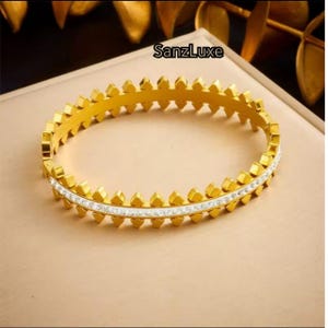 Może przedstawiać: Złota bransoletka o geometrycznym wzorze. Bransoletka posiada rząd przezroczystych, błyszczących kamieni i powtarzający się wzór trójkątnych kształtów. Widoczna jest nazwa marki "SanzLuxe". Ten element biżuterii to stylowy dodatek.