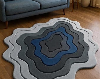 May include: An area rug featuring an irregular, wavy-edged shape with concentric layers in shades of light gray, medium gray, dark gray, and a central deep blue. The rug has a textured surface and a layered, topographical design.