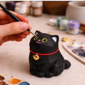 May include: A black cat figurine with a red collar and gold bell is being painted with a paintbrush. The cat has yellow eyes and white accents. The figurine is on a light-colored surface, with paint supplies in the background.