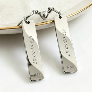 May include: Two silver-toned rectangular metal keychains connected by a small magnetic heart charm. Each keychain is engraved with the date 07.09.27, two small heart icons, and the initials M&L at the bottom. The metal has a polished, reflective finish.