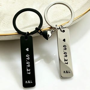 May include: Two metal keychains with circular rings, one black and one silver. Each features a rectangular pendant engraved with a heart symbol, the date 09.09.27, and the letters K&L. The pendants are connected by a small magnetic heart piece.