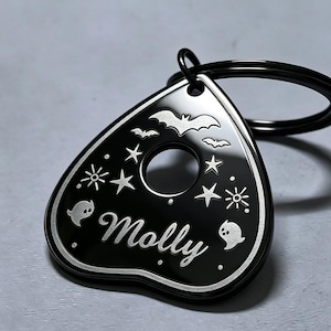 Könnte beinhalten: Ein schwarzer Schlüsselanhänger in Planchette-Form mit einer silberfarbenen Gravur von Fledermäusen, Sternen und zwei kleinen Geistern. Der Name Molly ist in Schreibschrift eingraviert, ergänzt durch eine kreisförmige Aussparung und einen Ring.