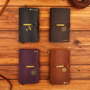 Op de afbeelding: Vier leren journals in zwart, bruin, paars en tan, elk met een unieke charme en gepersonaliseerde tekst. Het zwarte journal heeft een vliegtuigbedel en de datum "12.12.26". Het bruine journal heeft een kompasbedel en de naam "JASON". Het paarse journal heeft een klokbedel en het woord "DAD". Het tan journal heeft een boombedel en de naam "Sam".