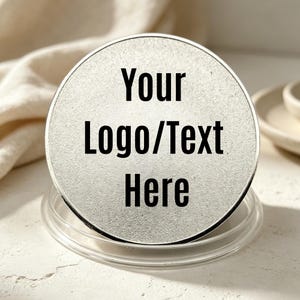 Può includere: Un disco metallico circolare color argento con una superficie testurizzata e il testo nero stampato "Your Logo/Text Here" centrato sul davanti. Il disco è posizionato verticalmente all'interno di una base di supporto circolare in plastica trasparente.