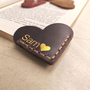 Könnte beinhalten: Ein herzförmiger Lesezeichen aus dunkelbraunem Leder mit dem Namen "Sam" in goldener Schrift und einem goldenen Herzen. Der Lesezeichen ist mit hellbraunem Faden genäht und liegt auf den Seiten eines Buches.