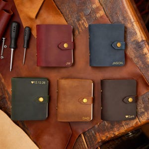 Puede incluir: Una colección de cuadernos de cuero personalizados en varios colores: burdeos, azul marino, verde oscuro, marrón y marrón oscuro. Cada cuaderno tiene un cierre de botón a presión dorado y está grabado con iniciales, nombres o una fecha. Se ven herramientas en el fondo.
