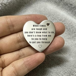 Könnte beinhalten: Ein silberfarbener, herzförmiger Metallanhänger mit einem kleinen ausgestanzten Herz oben rechts und einem eingravierten Herz unten. Der Text lautet: When times are tough and you don't know what to do, here's a hug from me to you to help to get you through.