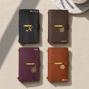 May include: Four leather journals in black, brown, purple, and orange. Each journal has a gold-coloured charm and a matching closure. The black journal has a plane charm and the date 12.12.26. The brown journal has the name Jason. The purple journal has the name Dad. The orange journal has the name Sam.