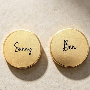 Peut inclure: Deux miroirs de poche circulaires de couleur dorée avec une surface texturée. Le miroir de gauche porte le nom Sunny en écriture noire, et le miroir de droite porte le nom Ben en écriture noire, tous deux centrés sur les couvercles métalliques.