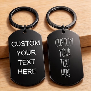 Könnte beinhalten: Zwei schwarze Metall-Schlüsselanhänger in Form von Erkennungsmarken mit runden Ringen. Der linke Anhänger zeigt den weißen Text CUSTOM YOUR TEXT HERE in fetter Schrift, der rechte denselben Text in einer dünnen, handschriftlichen Schrift.