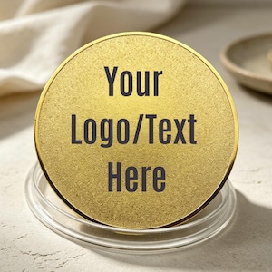 Könnte beinhalten: Eine kreisförmige, goldfarbene Scheibe mit strukturierter Oberfläche, auf deren Vorderseite der dunkelgraue Text "Your Logo/Text Here" zentriert ist. Die Scheibe steht aufrecht in einem runden, transparenten Kunststoffständer.