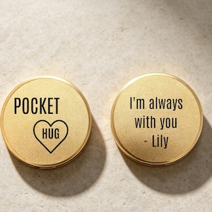 Könnte beinhalten: Zwei runde, goldfarbene Metallmünzen mit strukturierter Oberfläche. Die linke Münze zeigt den schwarzen Text POCKET HUG mit einem Herzsymbol. Die rechte Münze zeigt den schwarzen Text I'm always with you - Lily. Beide Objekte sind kreisförmig.