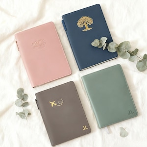 Könnte beinhalten: Vier ledergebundene Notizbücher in Rosa, Marineblau, Grau und Salbeigrün. Das marineblaue Notizbuch hat ein goldenes Baumdesign. Das graue Notizbuch hat eine goldene Flugzeuggrafik. Die Notizbücher in Salbeigrün und Grau haben goldene Initialen. Das rosa Notizbuch hat den Schriftzug "Not all those who wander are lost".