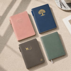 Könnte beinhalten: Vier ledergebundene Notizbücher in den Farben Rosa, Marineblau, Grau und Grün. Das rosa Notizbuch hat einen geprägten Text, das marineblaue Notizbuch zeigt ein goldenes Baumdesign, das graue Notizbuch hat eine goldene Flugzeuggrafik und das grüne Notizbuch hat ein weißes Band-Lesezeichen.